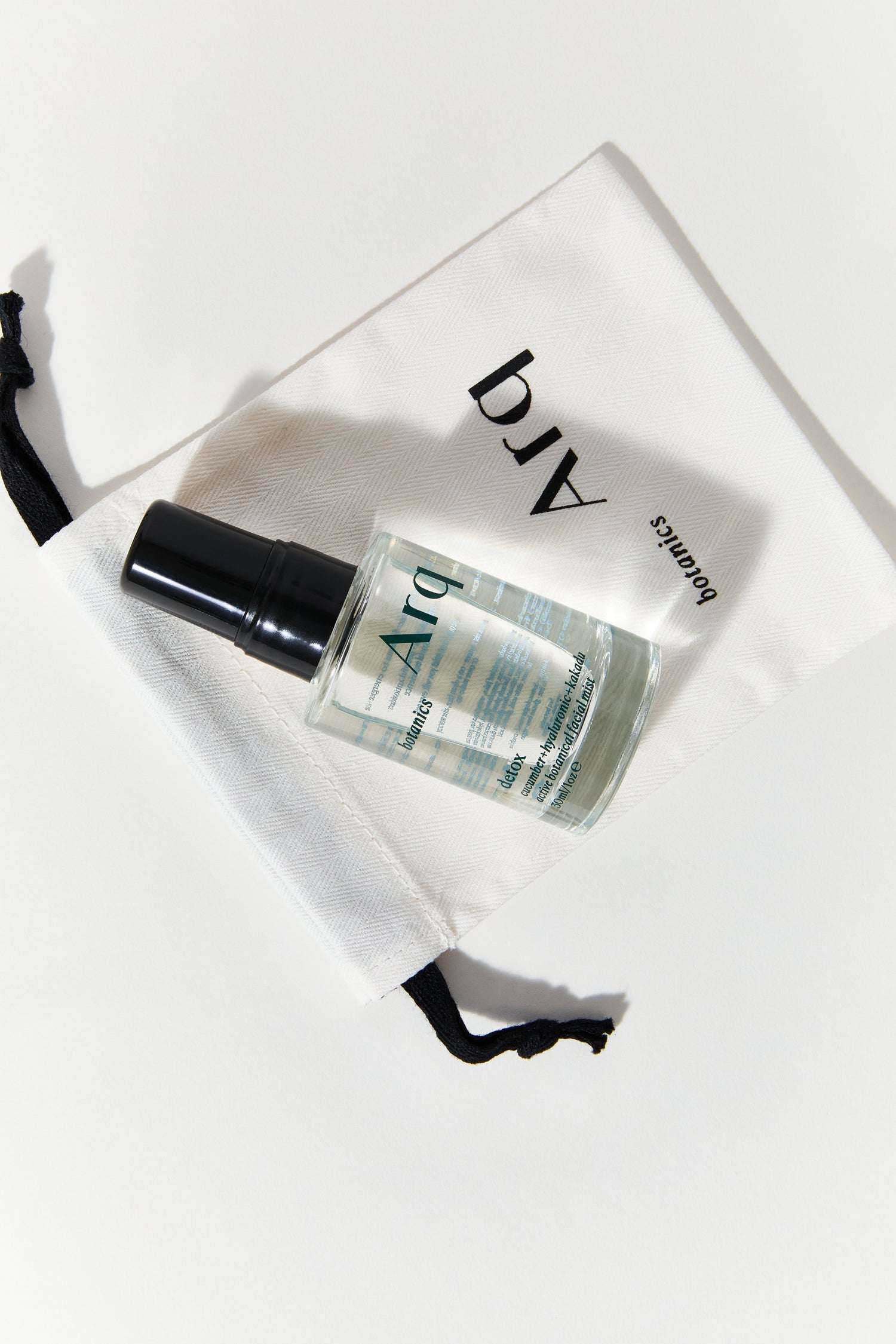 A small, clear spray bottle with a black cap, labeled "Arq Botanics Refresh Hyaluronic Kakadu." It's resting inside a white drawstring pouch with the "Arq Botanics" logo.
