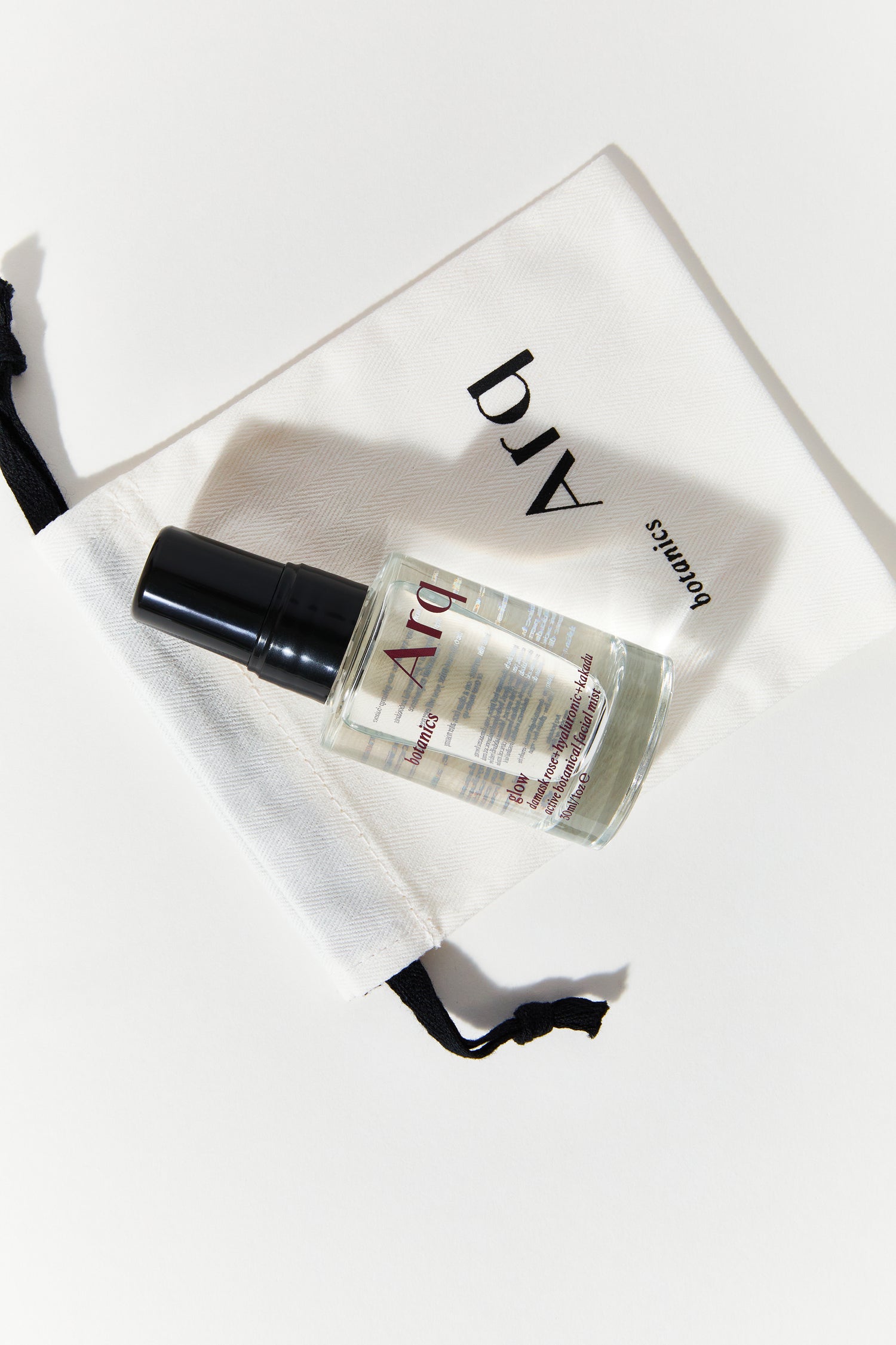 A small, clear spray bottle with a black cap, labeled "Arq Botanics Glow Rose Hyaluronic Kakadu." It's resting inside a white drawstring pouch with the "Arq Botanics" logo.