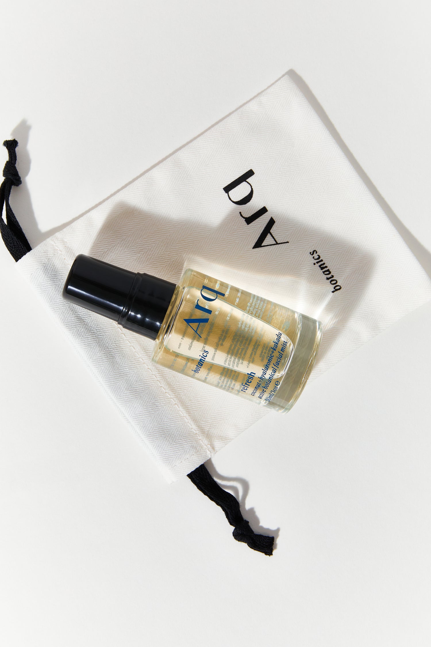A small, clear spray bottle with a black cap, labeled "Arq Botanics Refresh Hyaluronic Kakadu." It's resting inside a white drawstring pouch with the "Arq Botanics" logo.