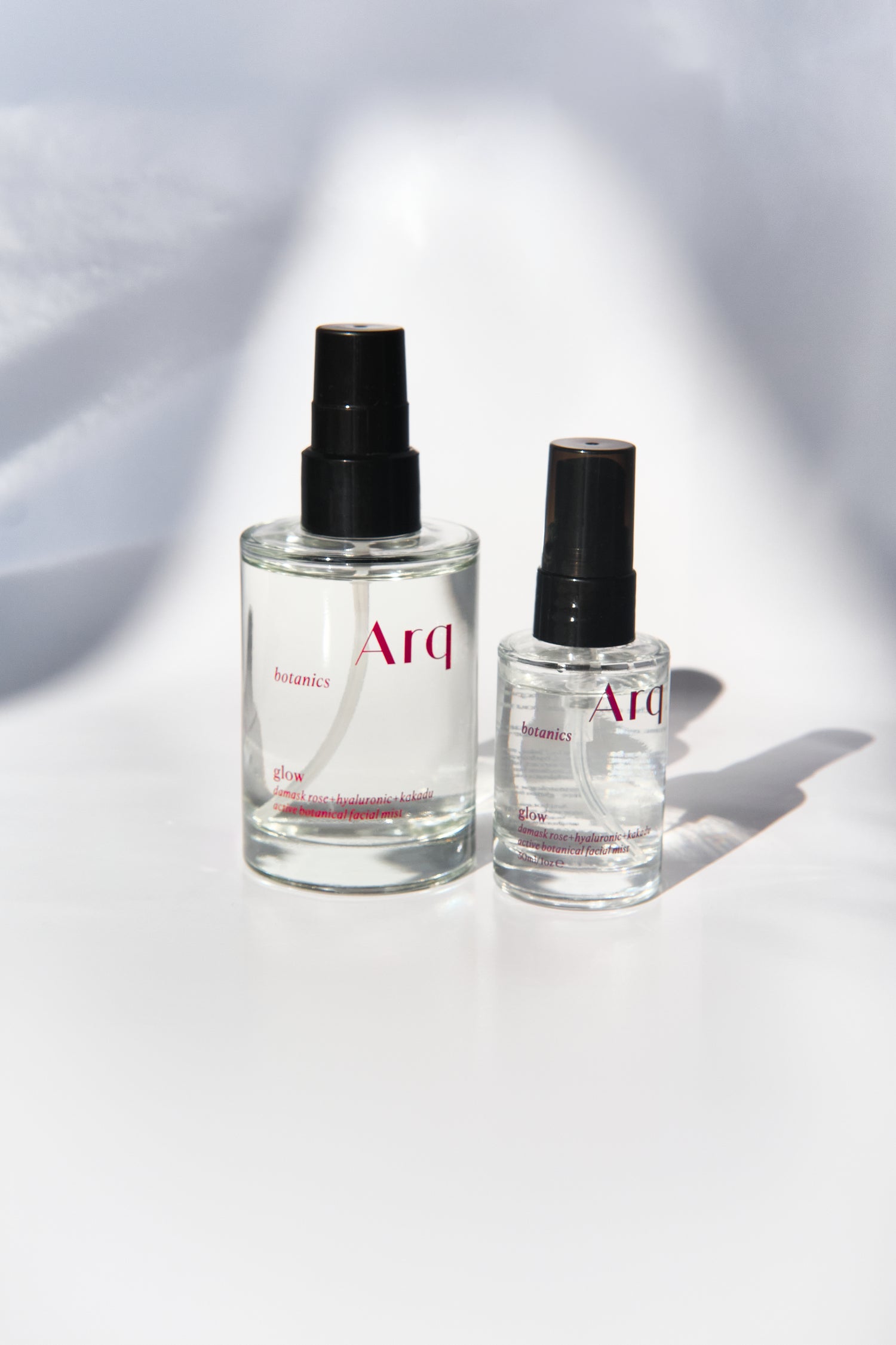 Two glass bottles of Arq Botanics Glow Rose Hyaluronic Kakadu Facial Mist. The larger bottle has a pump dispenser, and the smaller one has a spray nozzle. Both are resting on a white surface.