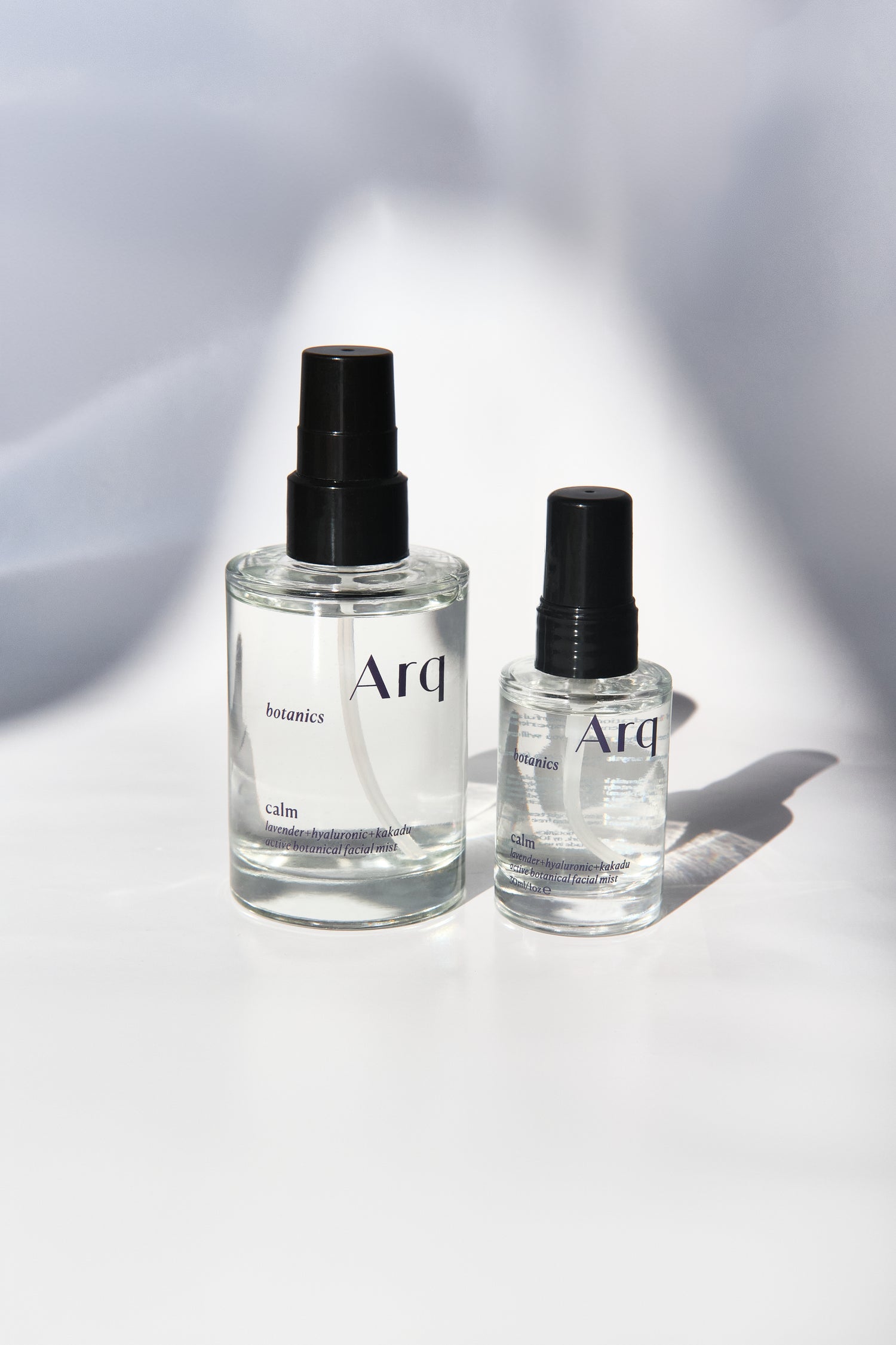 Two glass bottles of Arq Botanics Calm Lavender Hyaluronic Kakadu Facial Mist. The larger bottle has a pump dispenser, and the smaller one has a spray nozzle. Both are resting on a white surface.