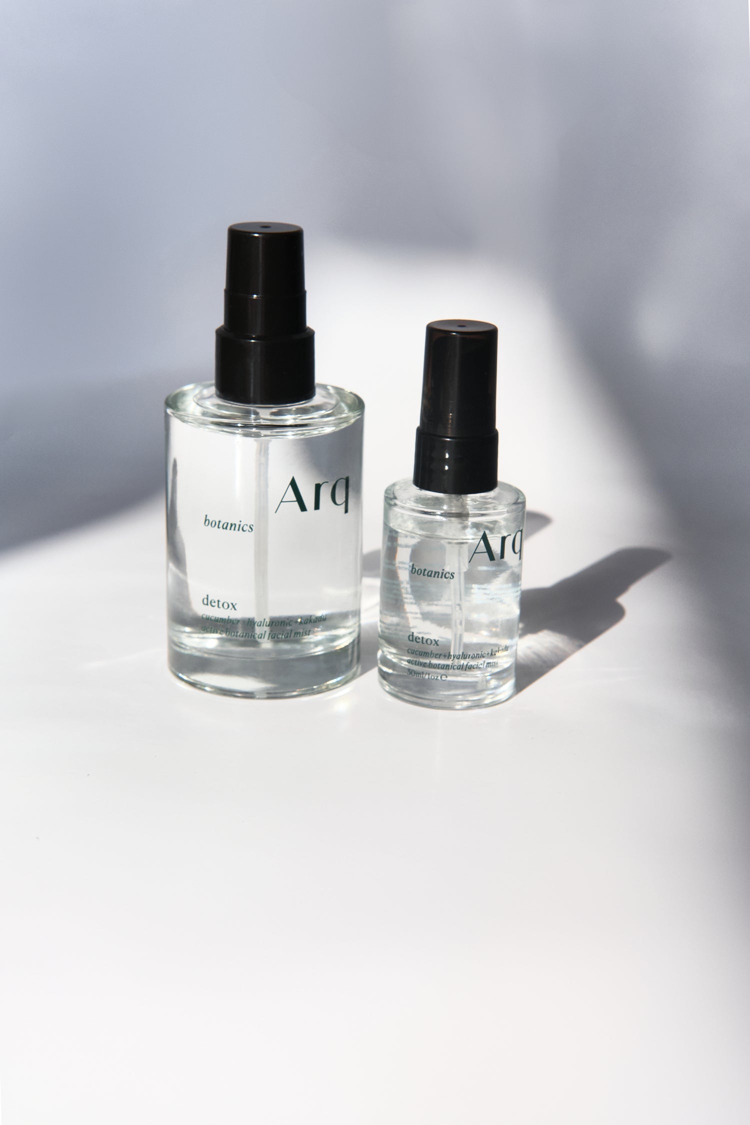 Two glass bottles of Arq Botanics Detox Cucumber Hyaluronic Facial Mist. The larger bottle has a pump dispenser, and the smaller one has a spray nozzle. Both are resting on a white surface.