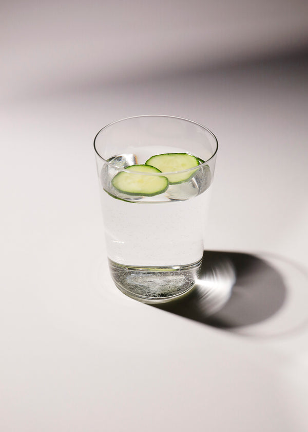 A tall glass filled with water and a few slices of cucumber, resting on a white surface.