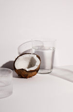A halved coconut with its white flesh exposed next to a clear glass of water on a wooden surface.