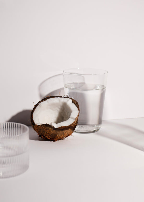 A halved coconut with its white flesh exposed next to a clear glass of water on a wooden surface.